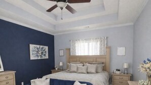 Ein Schlafzimmer mit einem Bett, einem blauen Sofa, einem Deckenventilator, einer Holzkommode und einem Fenster mit Vorhängen.