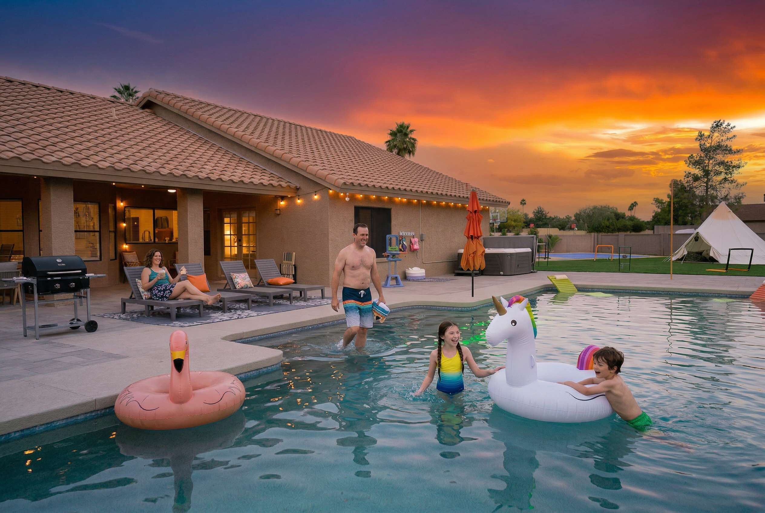 Una piscina con flotadores de unicornio inflables, una casa con tejado de baldosas y una puesta de sol al fondo.