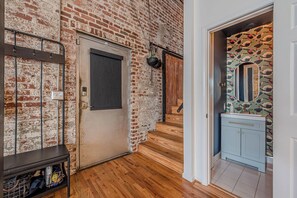 A hallway with a brick wall, a door, a bench, a wooden staircase, and a bathroom with a mirror and patterned wallpaper. - Historic Loft in Grant Park 3 less than 3 minute walk to MARTA Rail (Atlanta)