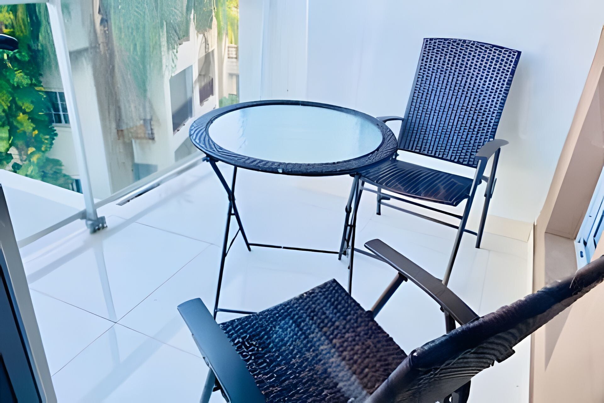 A balcony with a glass table and two chairs, a view of greenery outside, and a clear sky.
