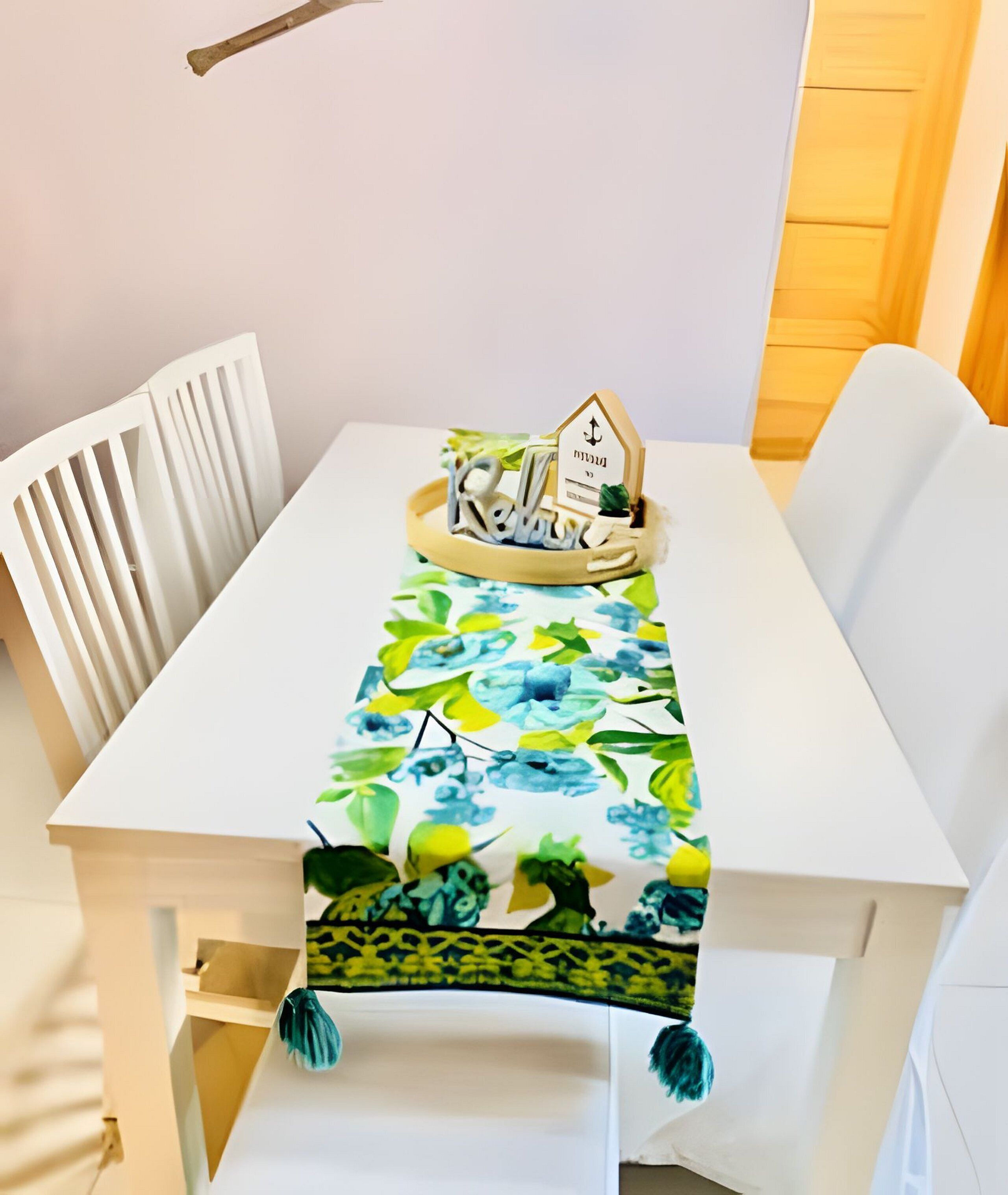 Ein weißer Tisch mit einer blumigen Tischdecke, einem kleinen, hausförmigen Schild und einem türkisfarbenen Quastenbespiel.