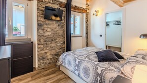 Une chambre avec un mur en pierre, des poutres en bois au plafond, un lit avec une literie à motifs, un téléviseur fixé au mur et une armoire.