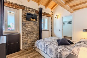 A bedroom with a stone wall, wooden ceiling beams, a bed with patterned bedding, a TV mounted on the wall, and a wardrobe. - The Best View Apartment Torno (Torno)