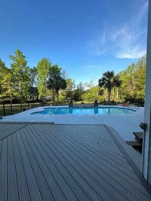Piscina com deck de madeira, cercada por árvores e uma vedação preta.