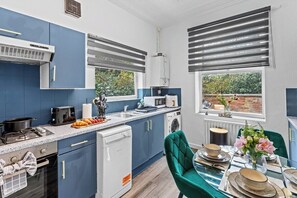 A compact kitchen with modern appliances, a dining area with a table set for two, and a view of greenery outside. - Winsick Residences Fallowfield up to 11 Guests (Manchester)