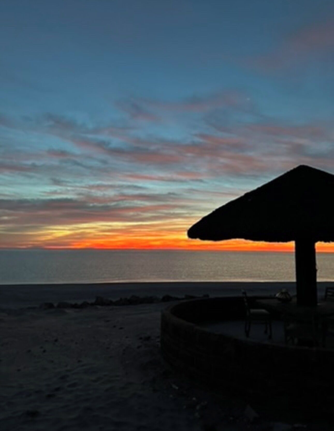 A beach scene with a thatched-roof structure, a sandy beach, and a sunset over the ocean.