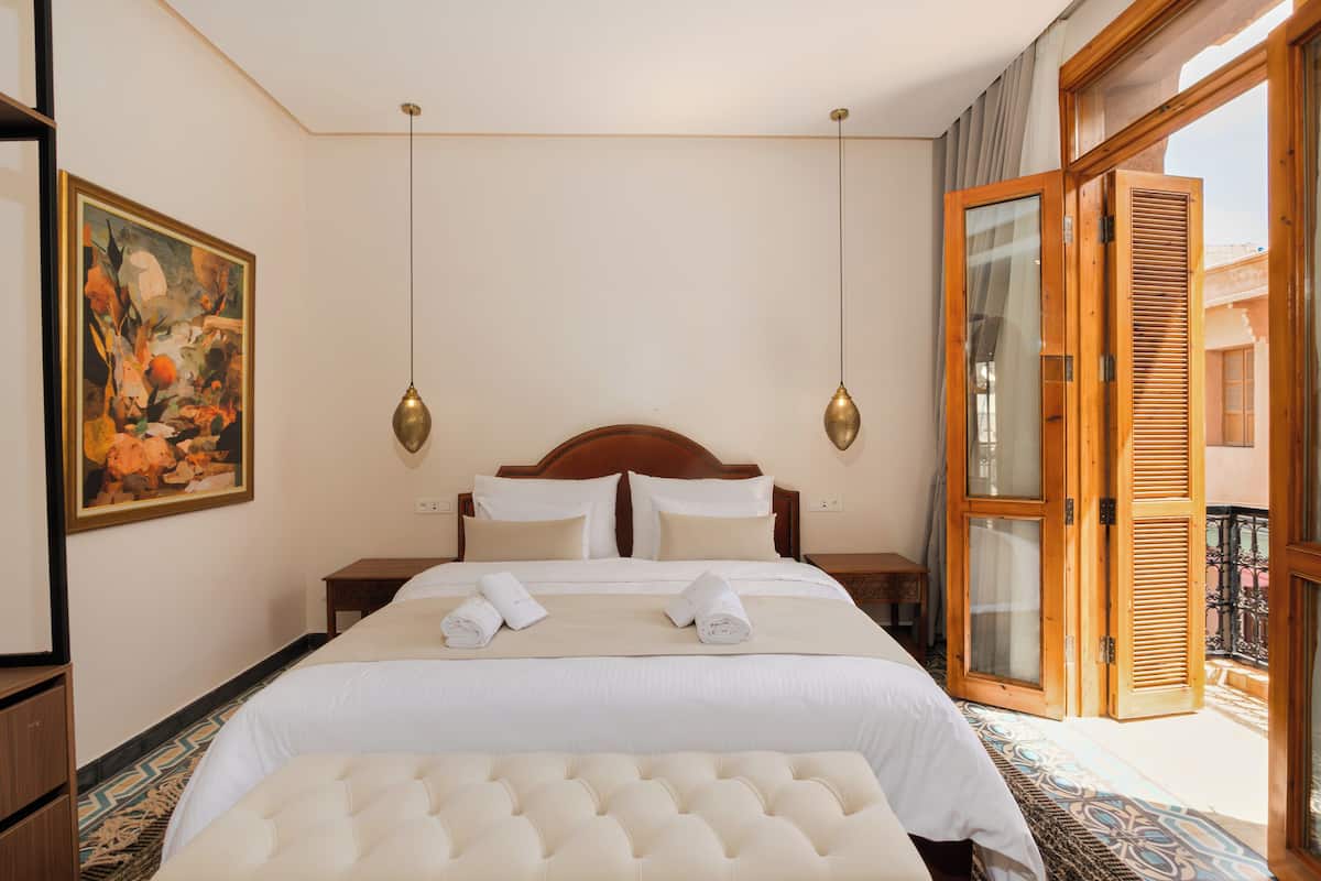 A hotel room with a large bed, a headboard, two bedside tables, a bench, a painting, and a balcony with wooden shutters.