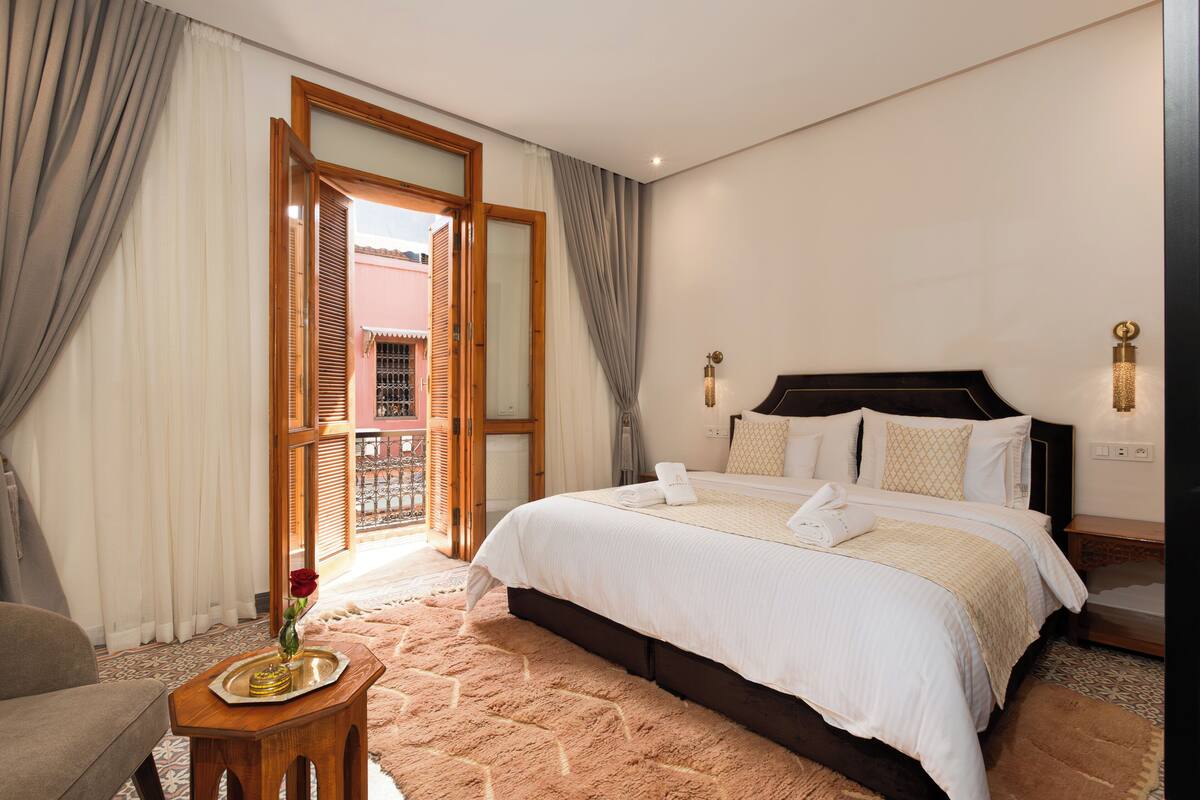 A bedroom with a bed, a small table with a vase and a tray, a chair, and a view through a door to an outdoor area.