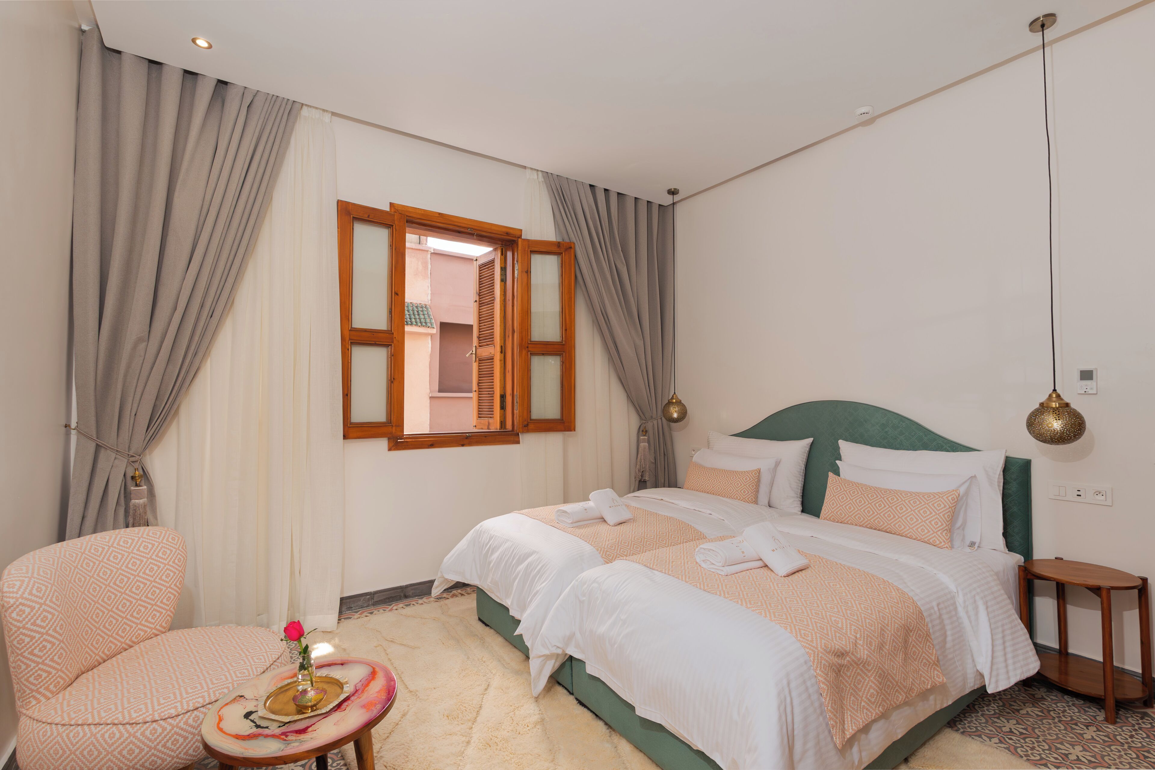A bedroom with a bed, a chair, a small table with a vase and a rose, and a window with curtains.