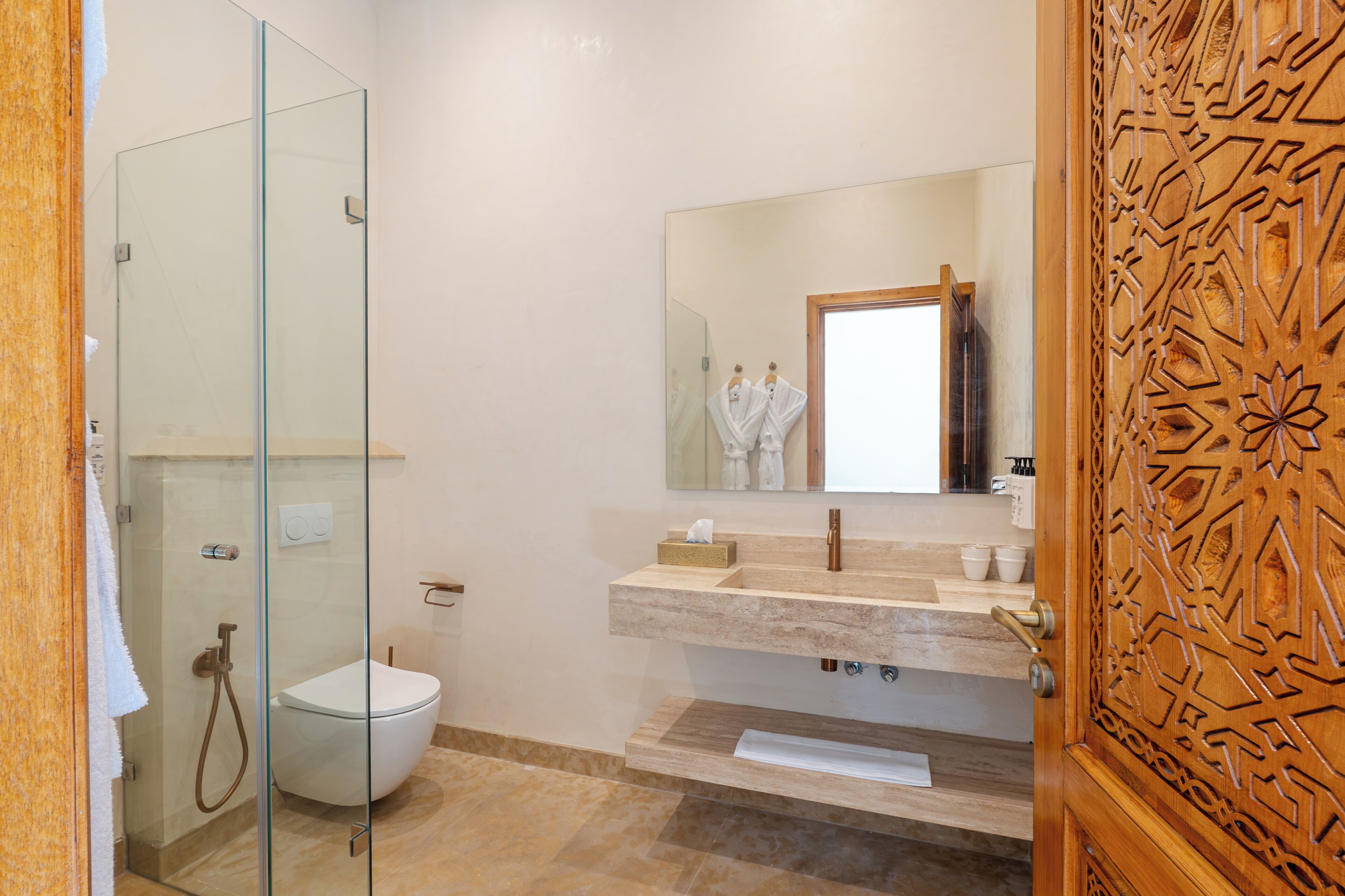 A modern bathroom with a glass shower enclosure, a white toilet, a vanity with a mirror, and a wooden door with intricate carvings.
