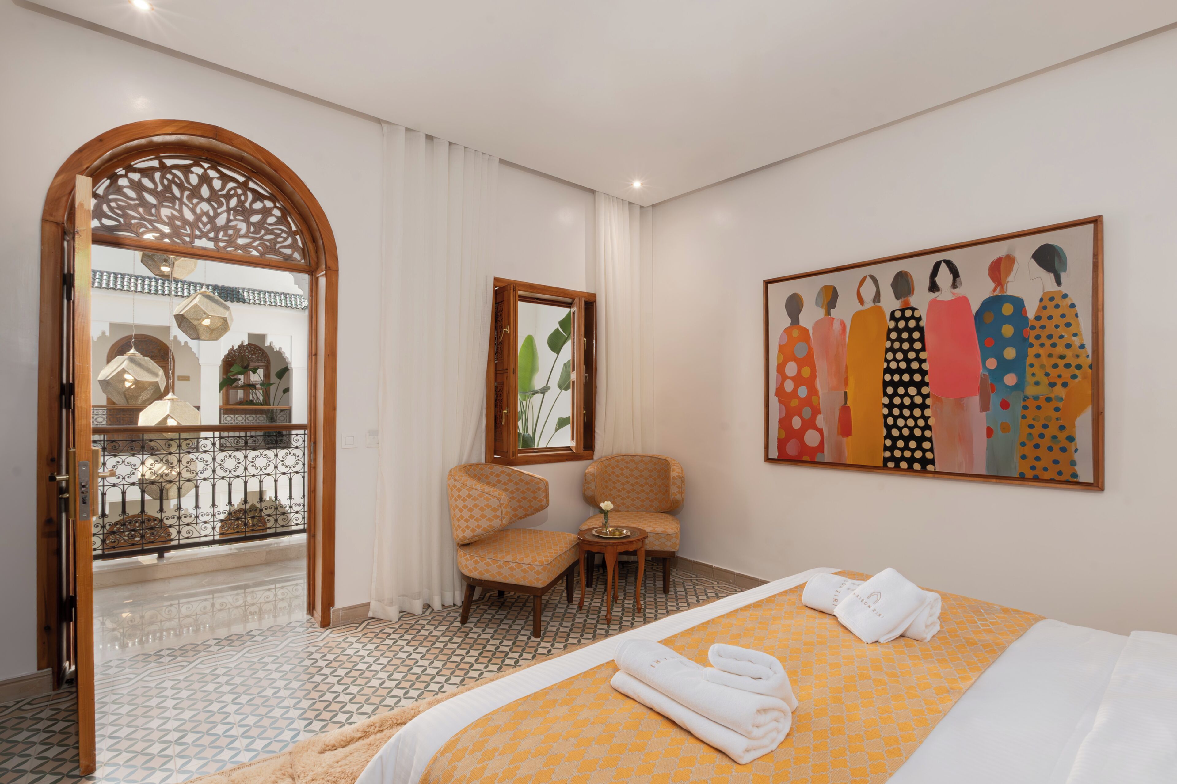 Une chambre d’hôtel avec un lit, deux chaises, une petite table, un tableau et une fenêtre décorative.