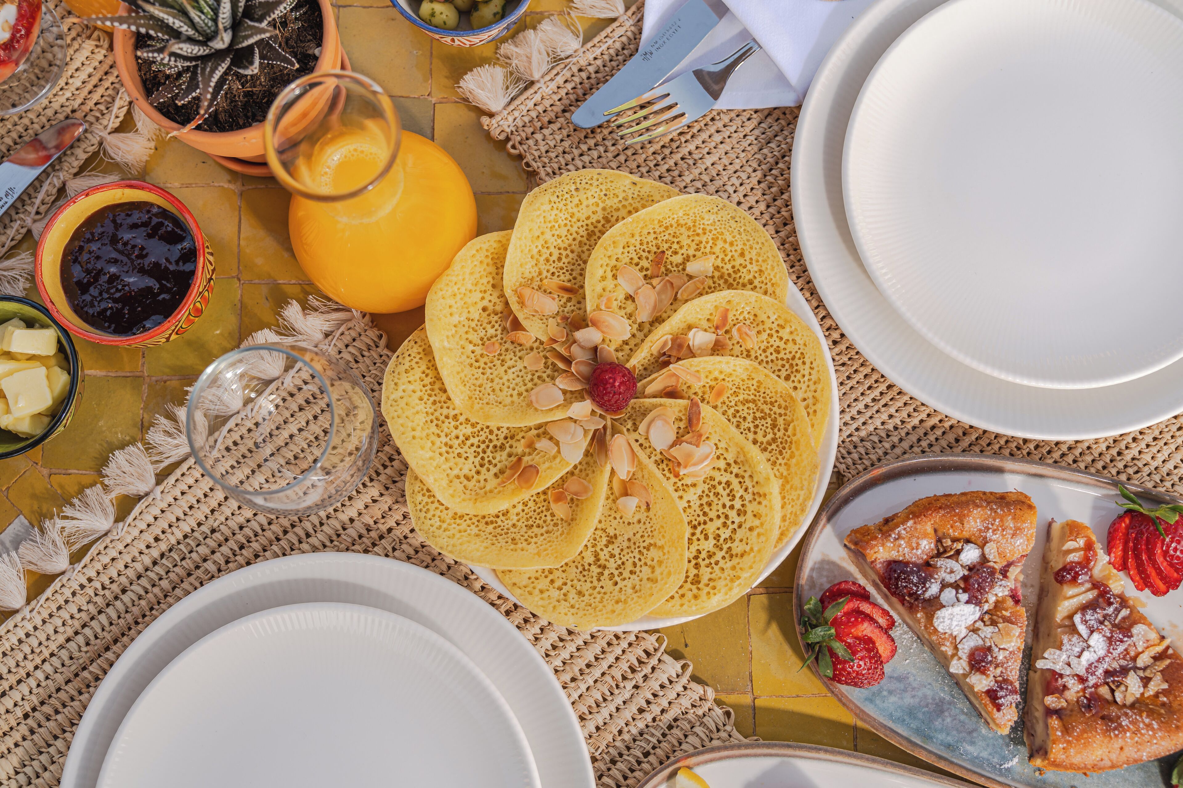 Une table dressée pour le petit-déjeuner avec des crêpes, des fraises coupées en tranches et un verre de jus d’orange.