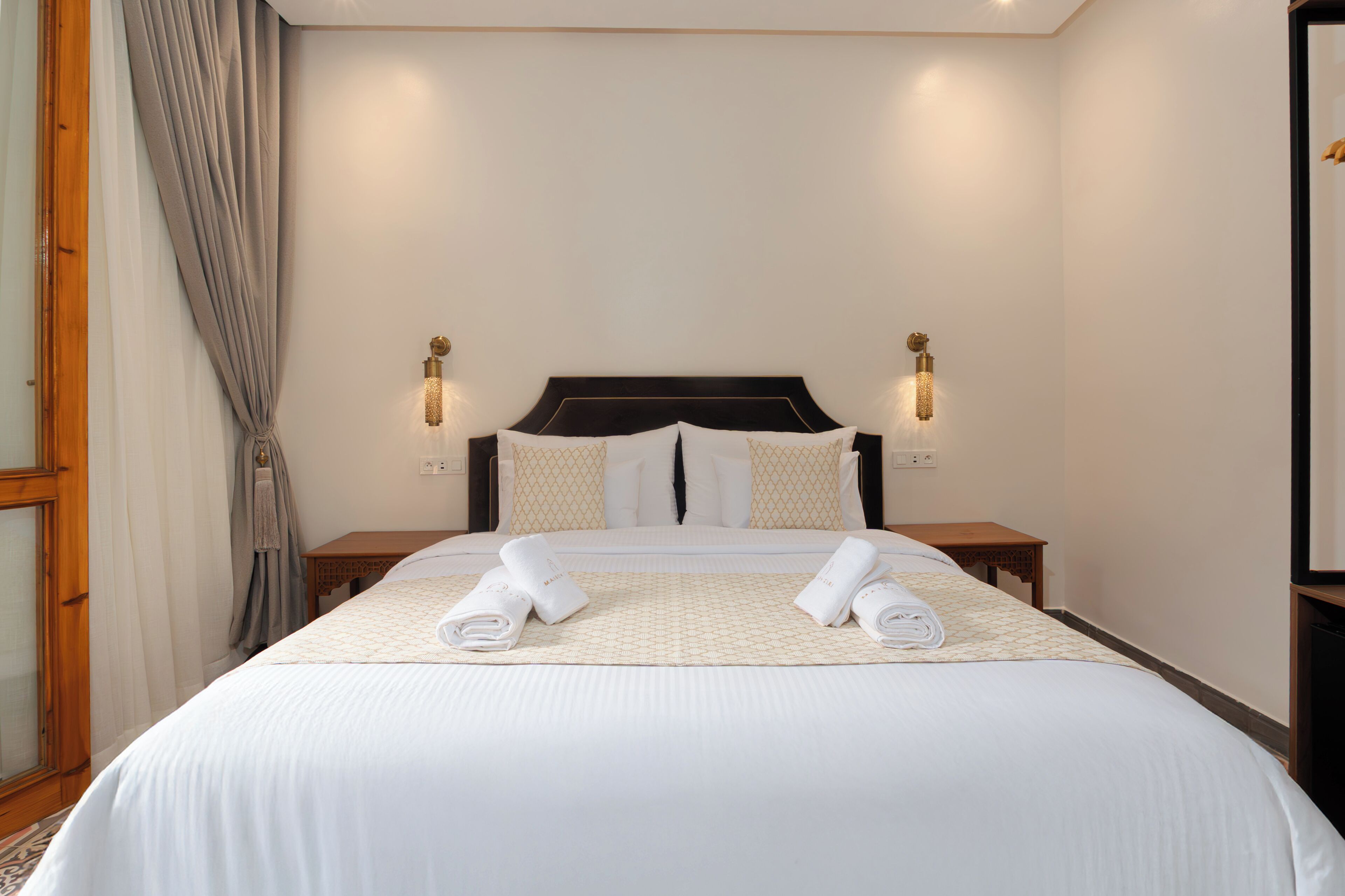 Un lit bien fait, avec du linge de lit et des serviettes blanches, une tête de lit sombre, deux tables de chevet avec des lampes et un plancher en bois.