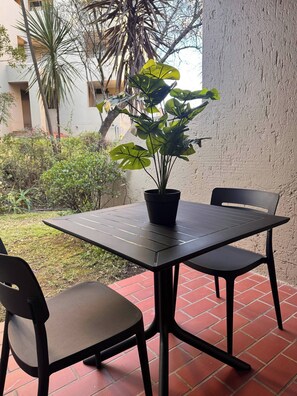 A patio with a table and two chairs, a potted plant, and a view of a building and greenery. - Stylish 1-bed in Bryanston (Johannesburg)