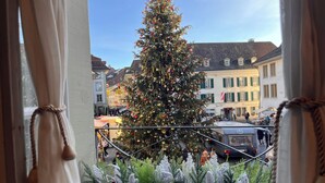 Blick durch ein Fenster mit Vorhängen, zeigt einen geschmückten Weihnachtsbaum und Gebäude im Hintergrund.