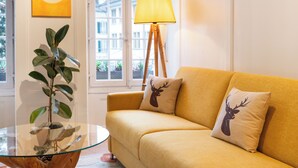Ein Zimmer mit einem Glastisch, einer Couch mit zwei Zierkissen, einer Stehlampe, einem Blumentopf und einem Fenster mit Blick auf Gebäude.
