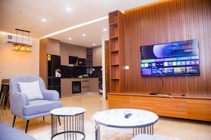 A modern living room with a flat-screen TV, a sofa, two side tables, and a kitchen area in the background. - Bravia Residency - Spring Properties (Accra)