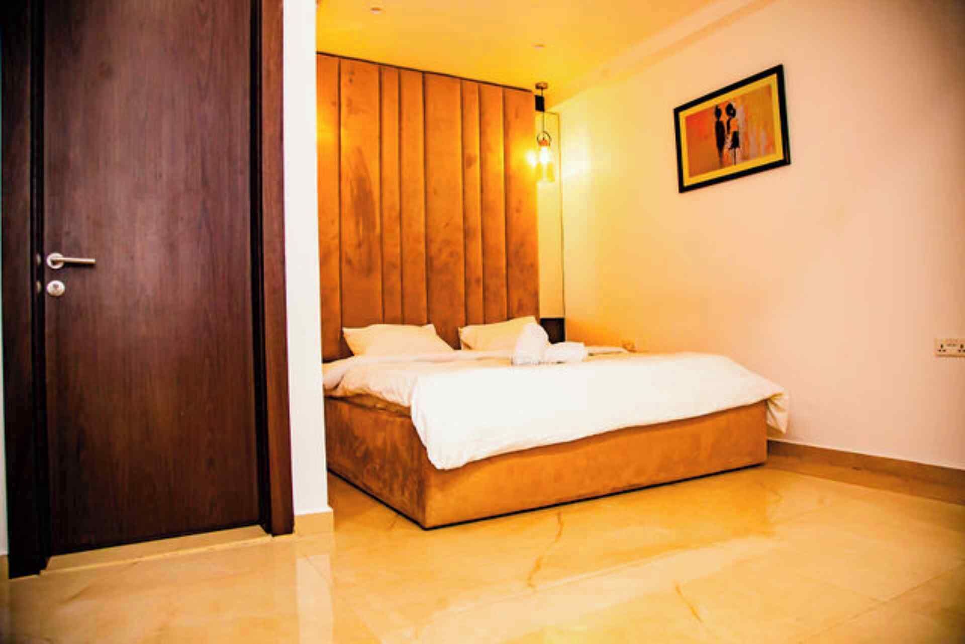 A bedroom with a wooden headboard, a bed with white bedding, a door, and a wall-mounted light fixture.