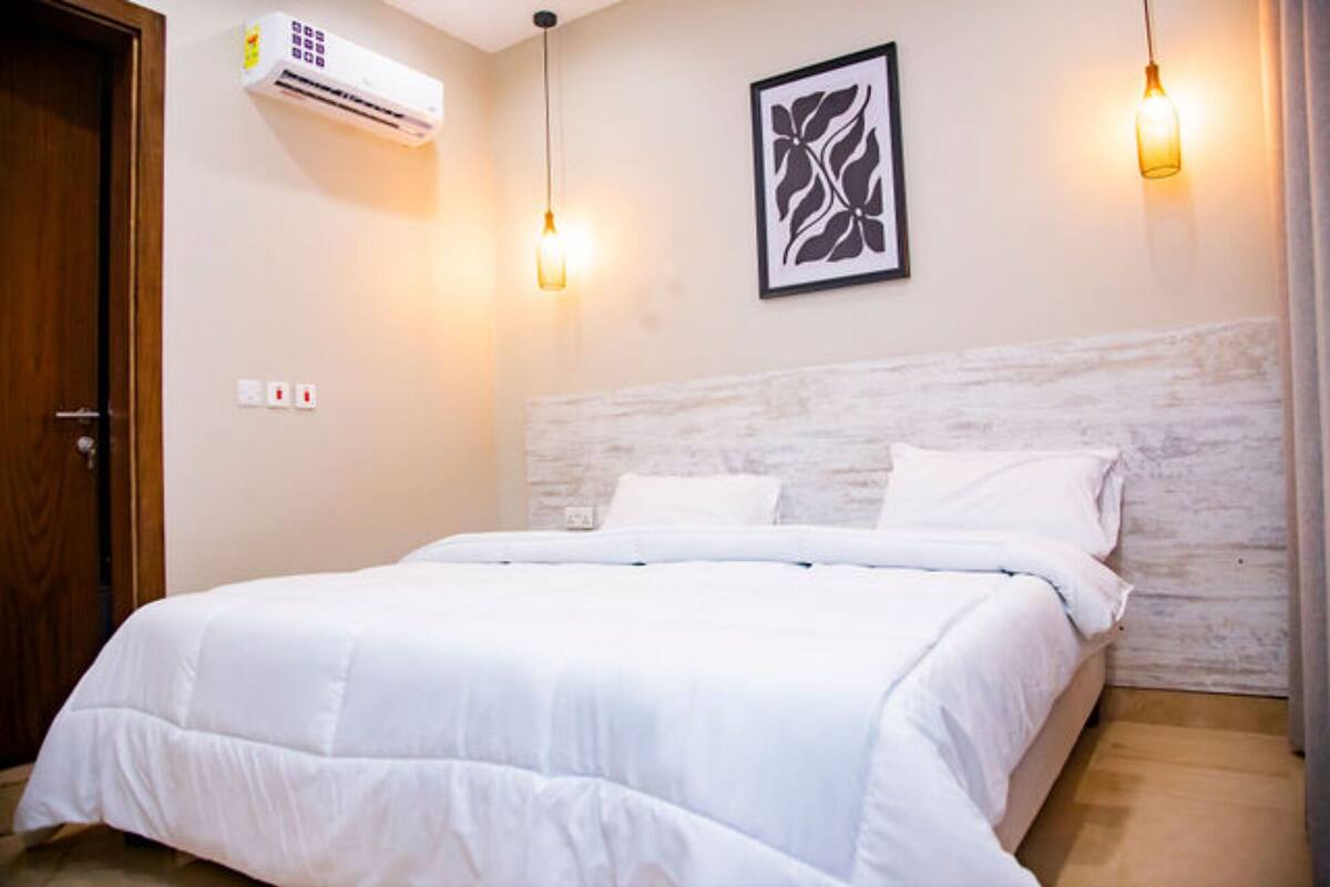 A hotel room with a bed, an air conditioning unit, a framed artwork, and two hanging light fixtures.