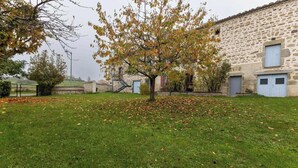 Ein Steinhaus mit einem Baum davor, einer grasbewachsenen Fläche und einem Zaun im Hintergrund.