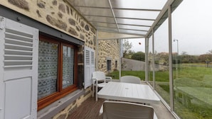 Eine überdachte Terrasse mit Tisch und Stühlen, ein Fenster mit Fensterläden und Blick auf eine Rasenfläche.