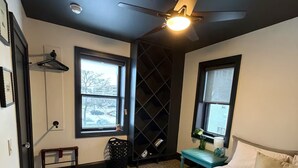 A bedroom with a bed, two windows, a ceiling fan, a wall-mounted shelf, and a door. - Urban Garden Loft - Historic Charm & Bold Design (Lansing)
