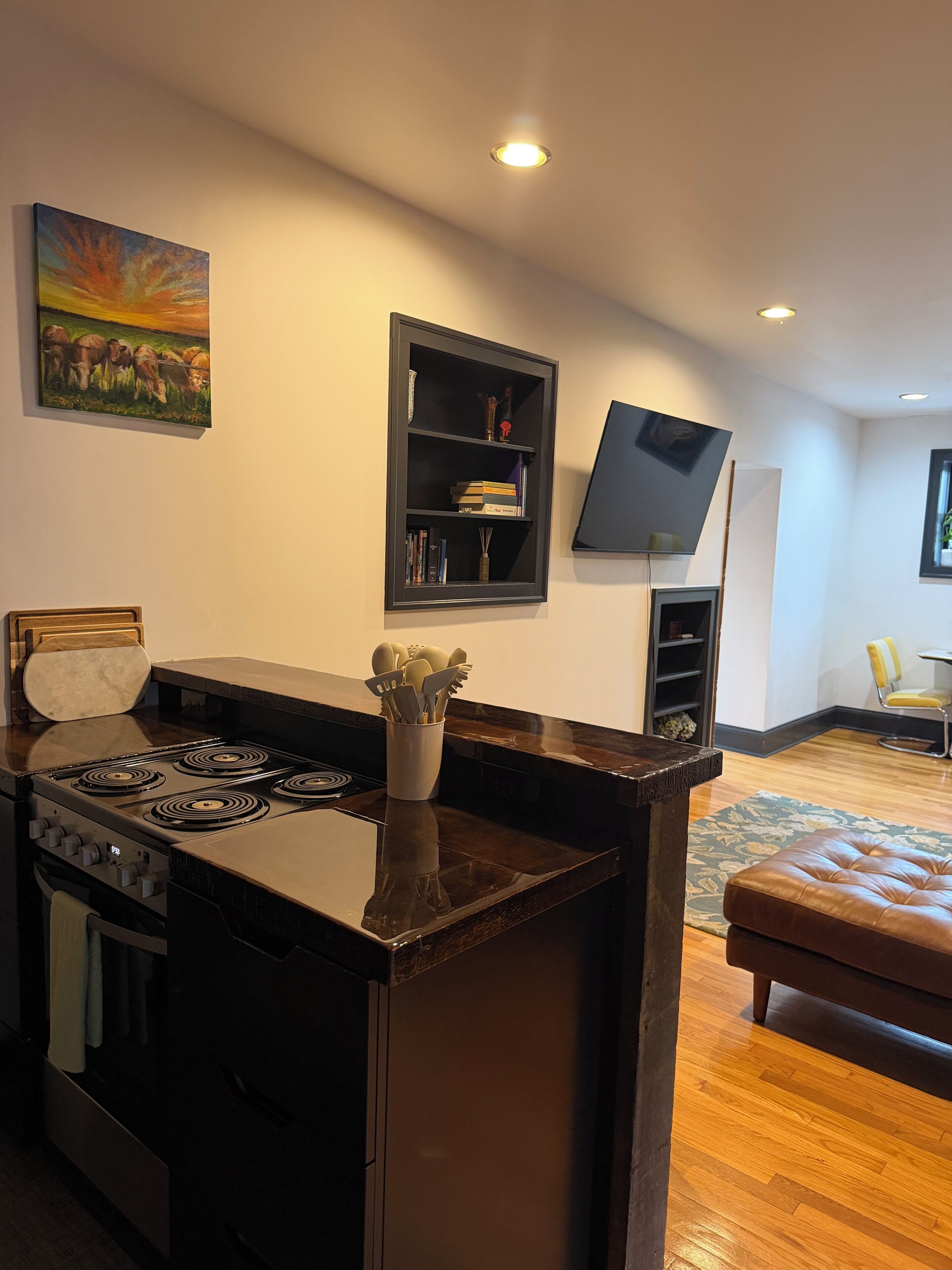 A modern kitchen with a gas stove, a dining table, and a bench. There is a bookshelf with books and decorative items.