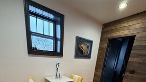A room with a wooden floor, a small table with two yellow chairs, a window with a view, and a door leading to another room. - Urban Garden Loft - Historic Charm & Bold Design (Lansing)