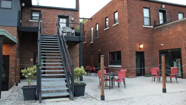 A brick building with a staircase leading to a balcony, outdoor seating area with red chairs, and a gravel ground. - Urban Garden Loft - Historic Charm & Bold Design (Lansing)