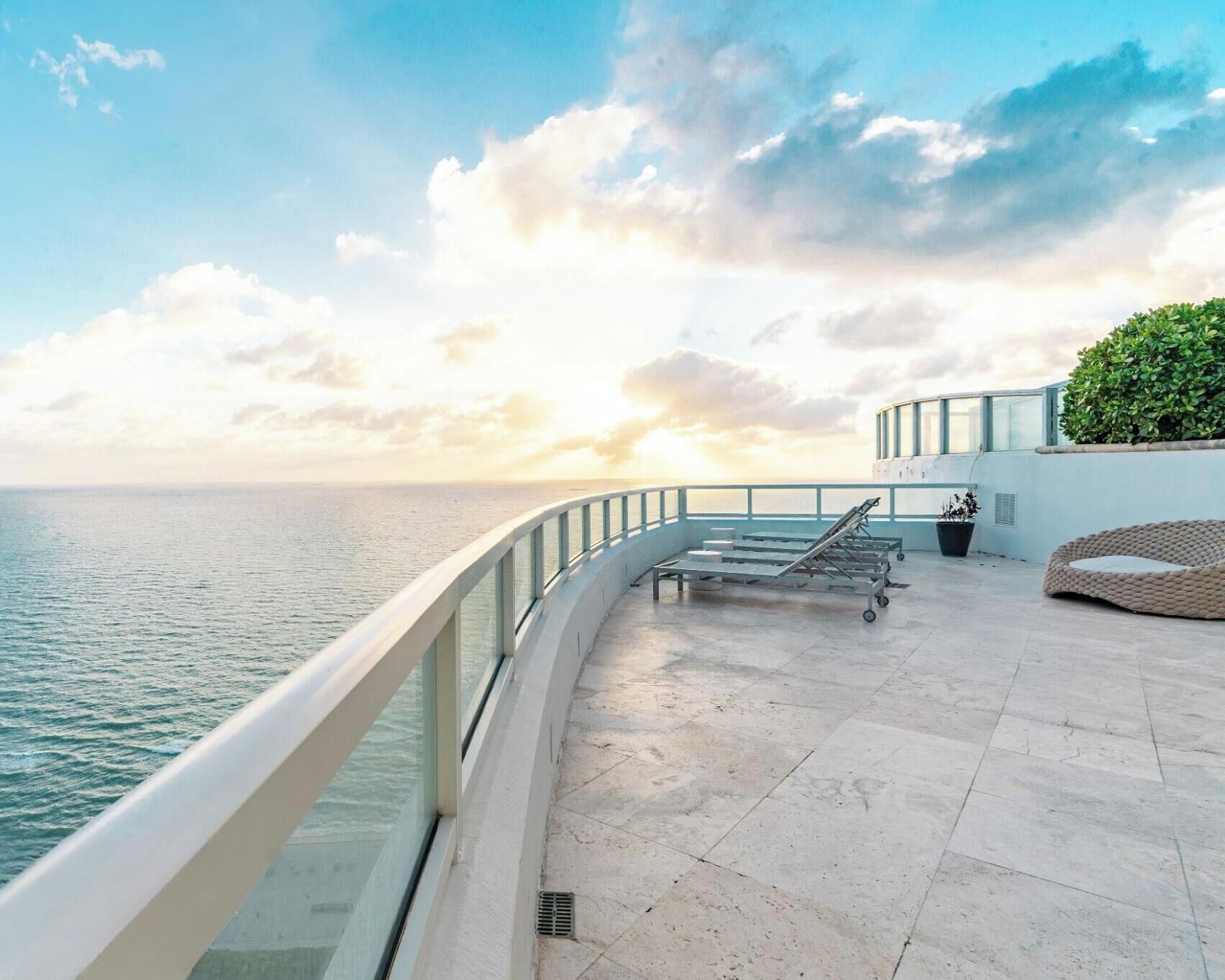 A sunlit terrace with a view of the sea, a railing, lounge chairs, and a potted plant.