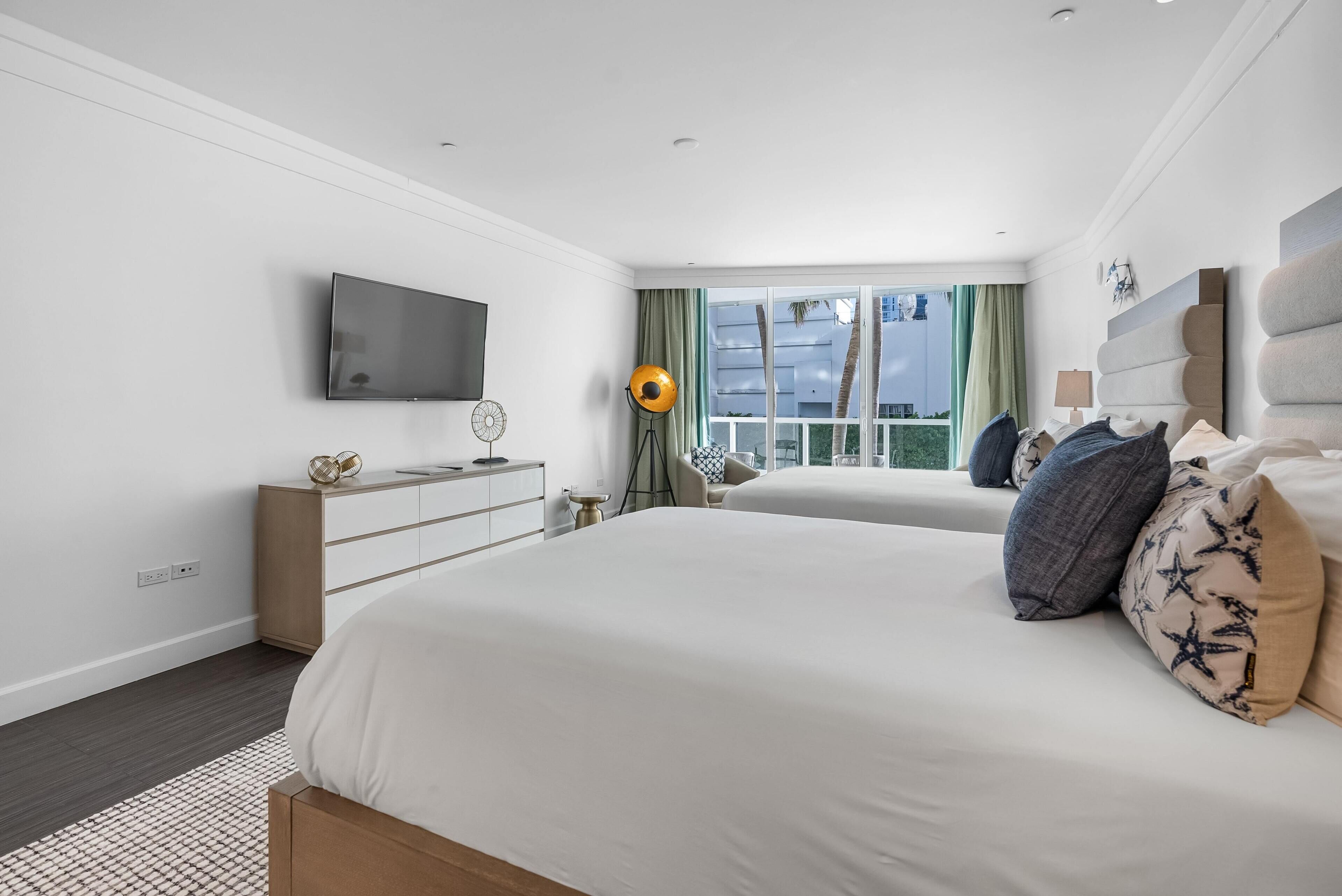 Ein modernes Hotelzimmer mit einem großen Bett, einem Flachbildfernseher, einem Schminktisch und Blick auf Palmen durch das Fenster.