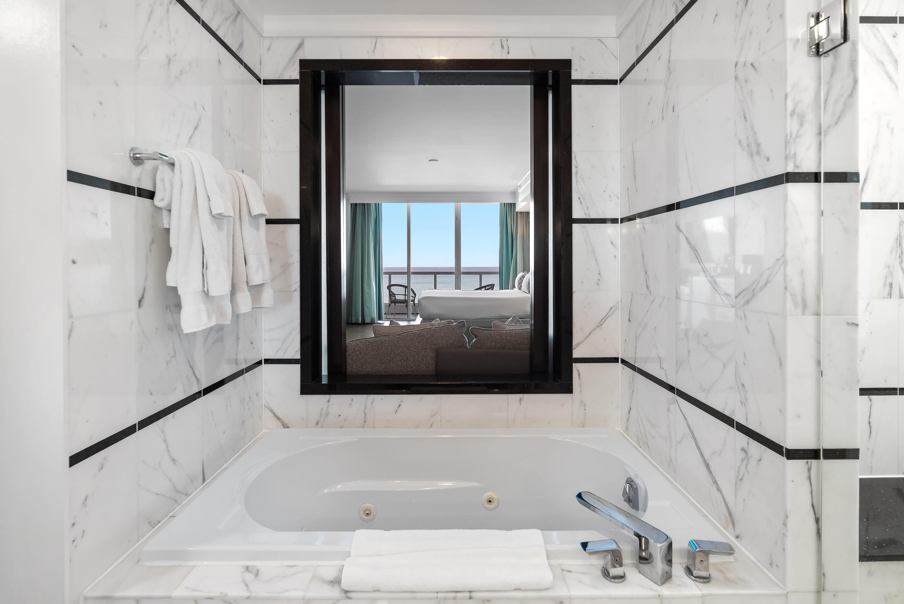 Ein modernes Badezimmer mit einer großzügigen Badewanne, einem Spiegel, der einen Schlafzimmer mit Balkon reflektiert, und Marmorwänden.