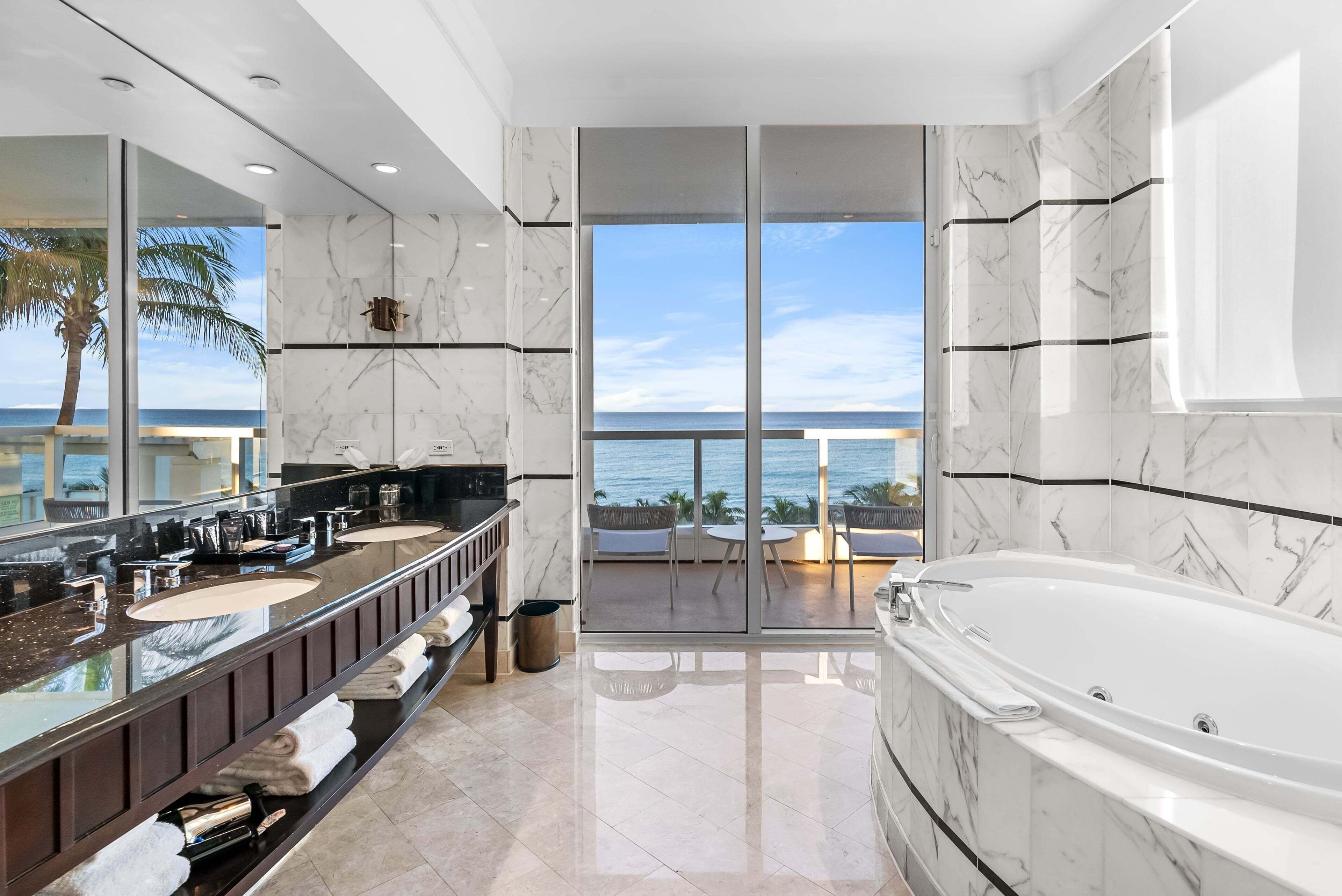 A modern bathroom with a large mirror, two sinks, a bathtub, and a view of the ocean.