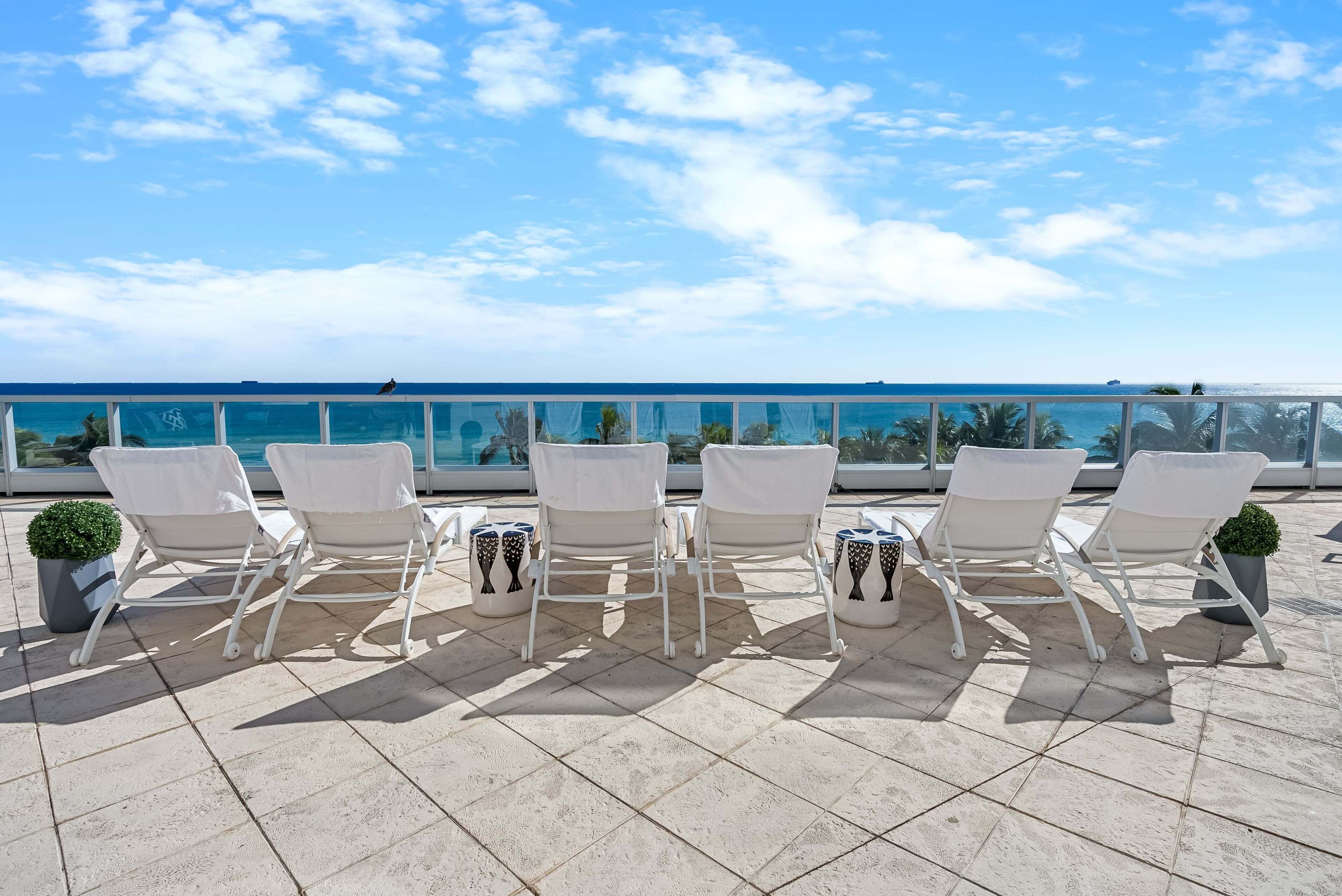 Une terrasse sur le toit, avec des chaises blanches et des plantes en pot, d’où l’on peut admirer un ciel bleu dégagé et l’océan.