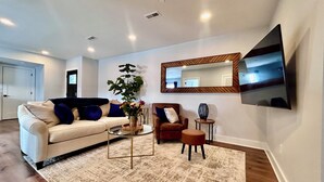 Una sala de estar con sofá, sillón, mesa de centro y televisor montado en la pared.