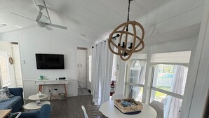 A modern living room with a flat-screen TV, a ceiling fan, a dining area with a round table and chairs, and a large window with curtains.