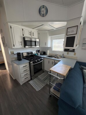 Una kitchenette compacta con estufa, microondas y lavamanos, un sofá azul y un reloj en la pared.