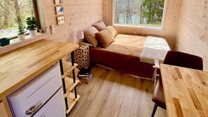 Een gezellig chalet met een bed, een kleine tafel, een koelkast, een raam met uitzicht op bomen en een wandgemonteerde airconditioner.