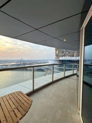 A balcony with a view of a river and cityscape, a wooden bench, and a glass railing. - Hotel apartment in Hilton Nile Maadi with a charming Nile and pyramids view (Cairo Governorate)
