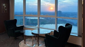 Quarto de hotel moderno com uma grande janela que oferece vista para a cidade, uma mesa de vidro redonda, duas poltronas e um objeto decorativo na parede.