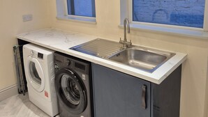 Une petite buanderie équipée d’un lave-linge, d’un sèche-linge et d’un évier. Deux fenêtres laissent entrer la lumière naturelle.