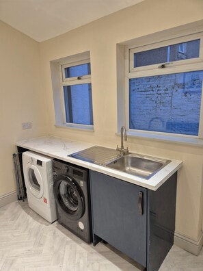 A compact laundry room with a washing machine, dryer, and a sink. There are two windows providing natural light. - Cosy Spacious Coastal Stay (South Shields)