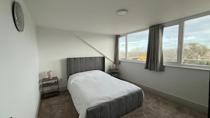 A bedroom with a bed, a bedside table, a lamp, a clock, and a window with curtains. - Cosy Spacious Coastal Stay (South Shields)