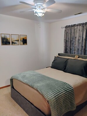 Un dormitorio con cama, ventilador de techo, ventana con cortinas y tres cuadros de paisajes en la pared.