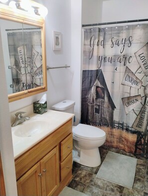 Un baño con tocador de madera, un lavamanos blanco, un inodoro y una cortina de ducha con texto inspirador.