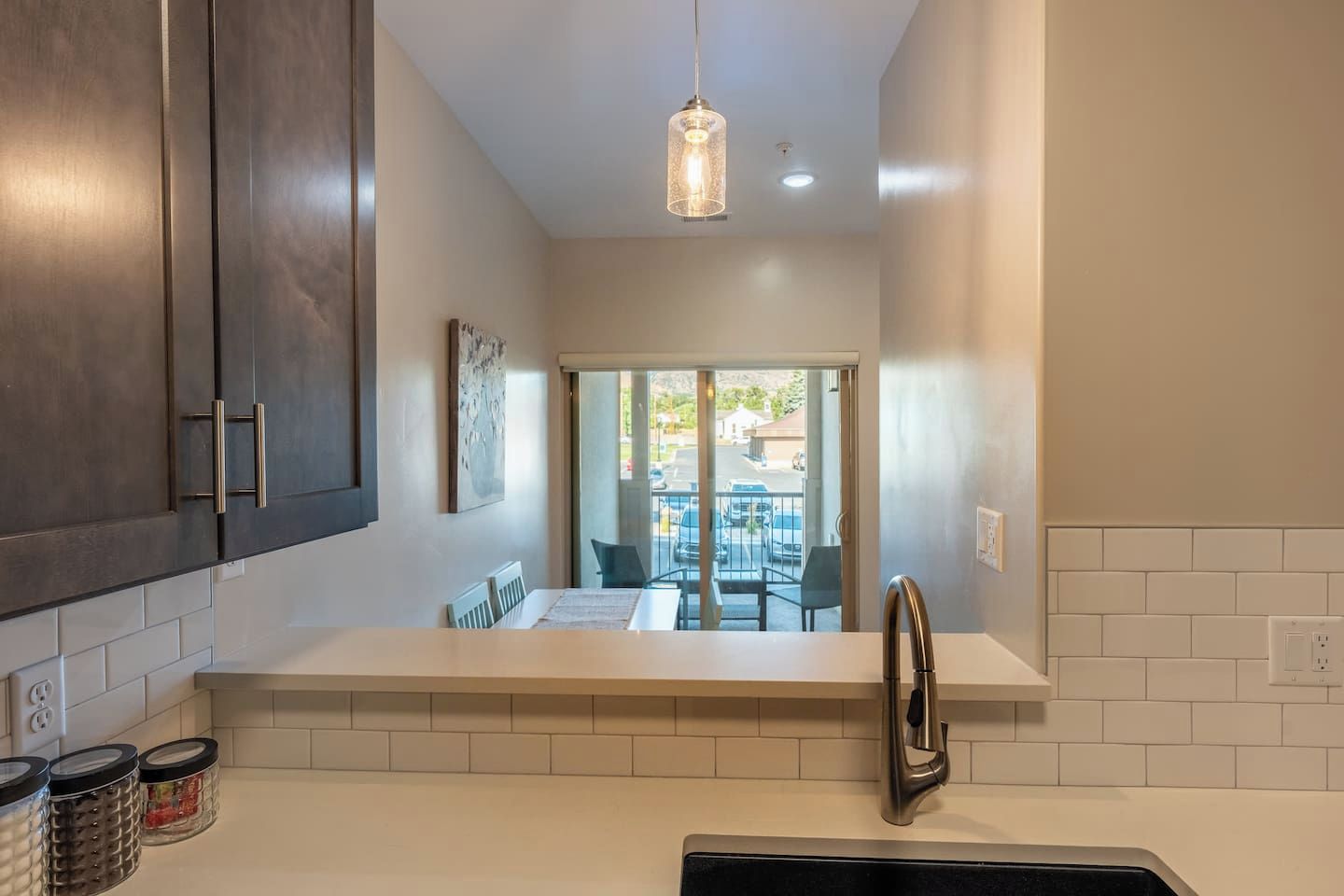 Une cuisine moderne avec des meubles de couleur sombre, un plan de travail blanc, un luminaire suspendu et une vue sur une terrasse extérieure.