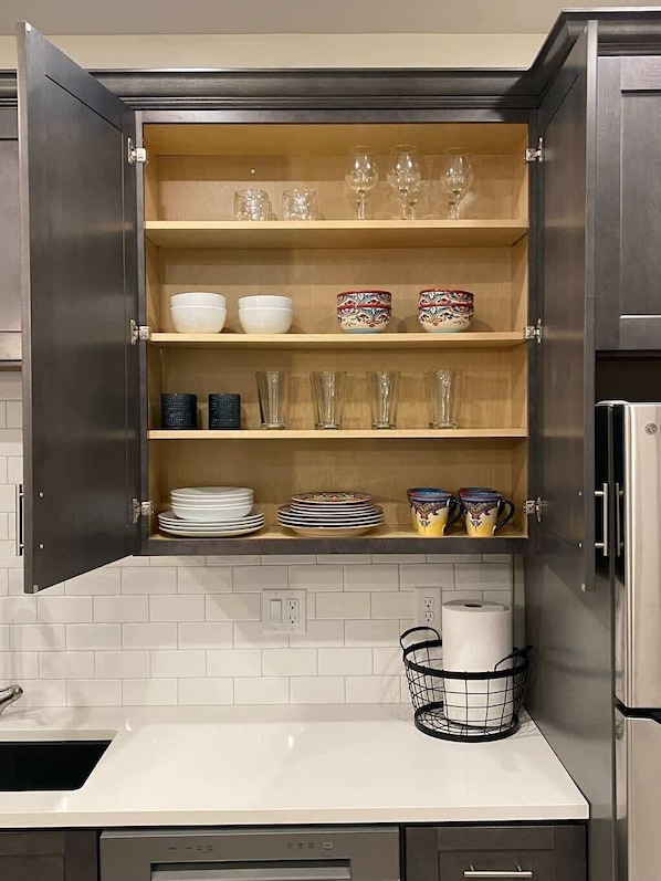 A kitchen with a cabinet containing bowls, plates, glasses, and a basket with paper towels.
