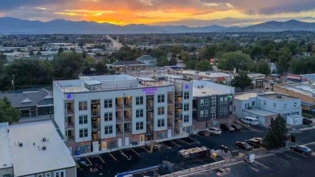 A multi-story building with a parking lot in front, surrounded by other buildings and mountains in the background. - Cozy Main St Stay | King Bed + Loft Bed | Sleeps 3 (Pleasant Grove)