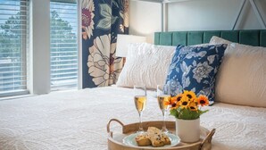A neatly made bed with a tray of food and drinks, a vase of flowers, and a view of the outdoors through a window with blinds. - Cozy Main St Stay | King Bed + Loft Bed | Sleeps 3 (Pleasant Grove)