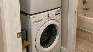 A stacked washer and dryer unit in a bathroom with a bathtub visible in the background. - Cozy Main St Stay | King Bed + Loft Bed | Sleeps 3 (Pleasant Grove)